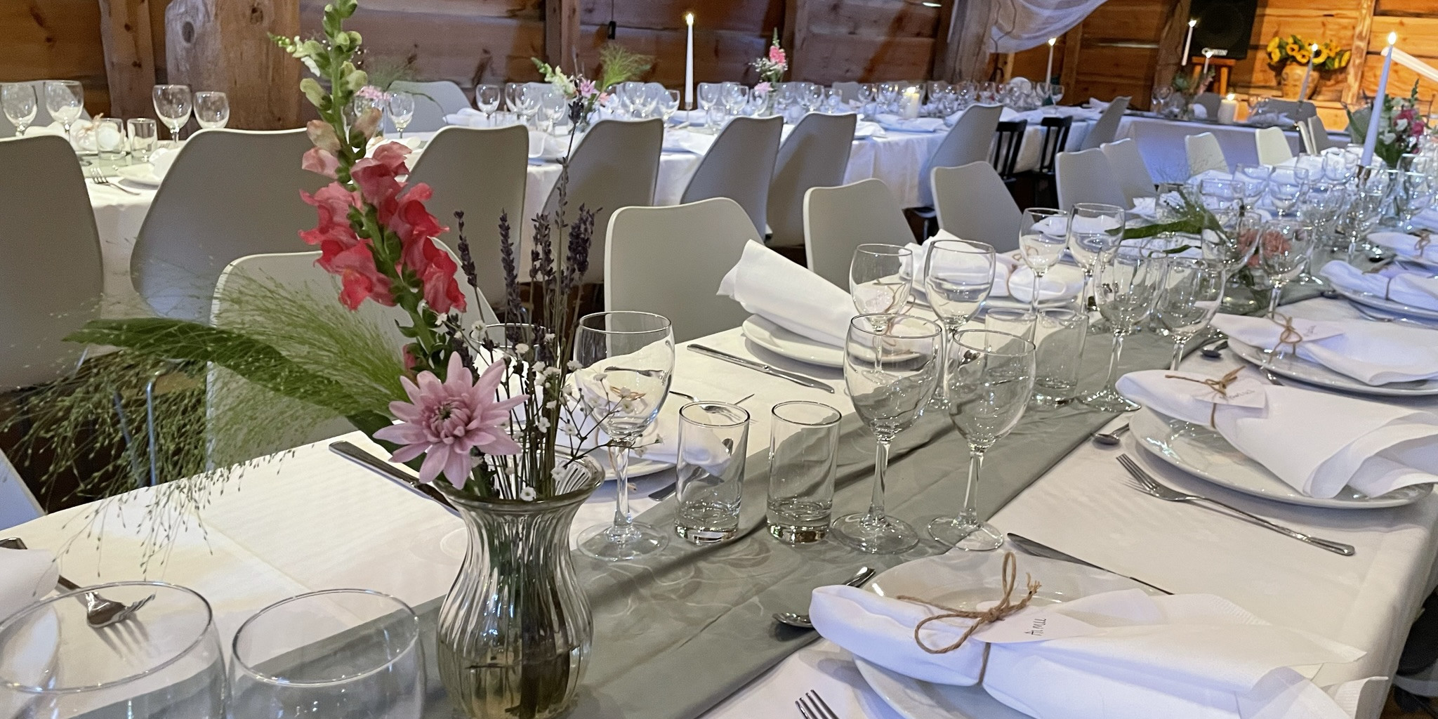 Utsnitt av odekorerte langbord til bryllaup. Borda er pynta med kvite dukar, stearinlys, serviettar med namnelappar og blomar. I bakgrunnen kvit tyll som er festa i tak og lys.