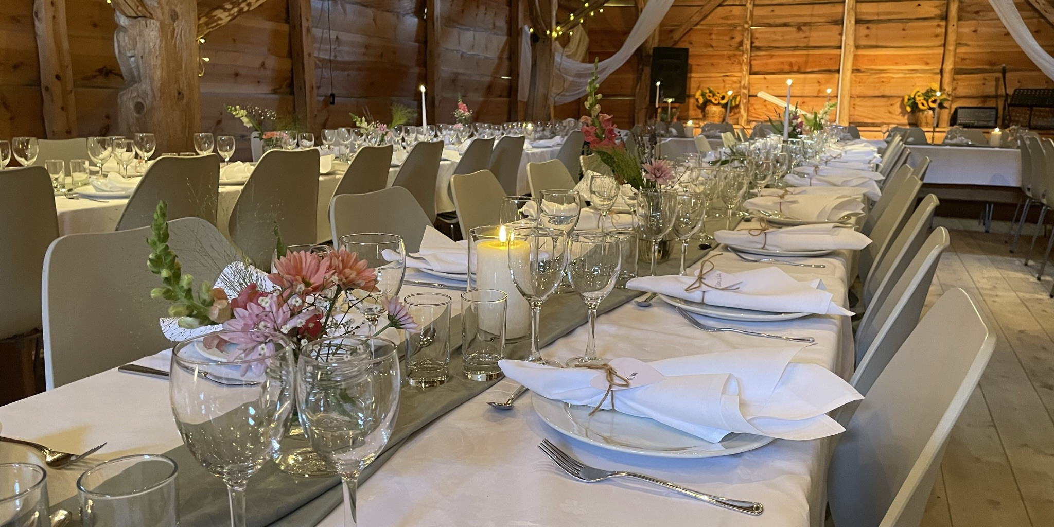 Utsnitt av odekorerte langbord til bryllaup. Borda er pynta med kvite dukar, stearinlys, serviettar med namnelappar og blomar. I bakgrunnen kvit tyll som er festa i tak og lys.